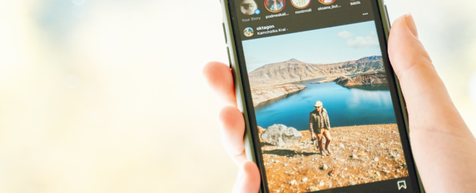 Hand holding a smartphone displaying an Instagram feed, featuring a scenic outdoor image of a person standing by a lake with mountains in the background