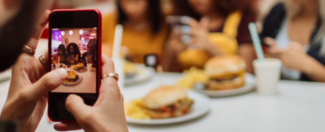 Person taking a photo of a group of friends at a table enjoying burgers and fries, highlighting the power of capturing real-life moments for social media storytelling